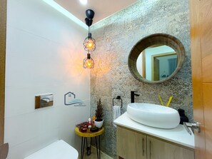 Ein Badezimmer mit einem weißen Waschbecken, einem runden Spiegel, einer dekorativen Leuchte und einem kleinen Tisch mit Toilettenartikeln.