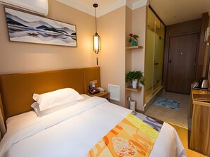 A hotel room with a bed, a bedside table with a phone and a lamp, a painting on the wall, a potted plant, and a wooden door. - Shell Zhengzhou Zhongyuan Fota Hotel (Zhengzhou)