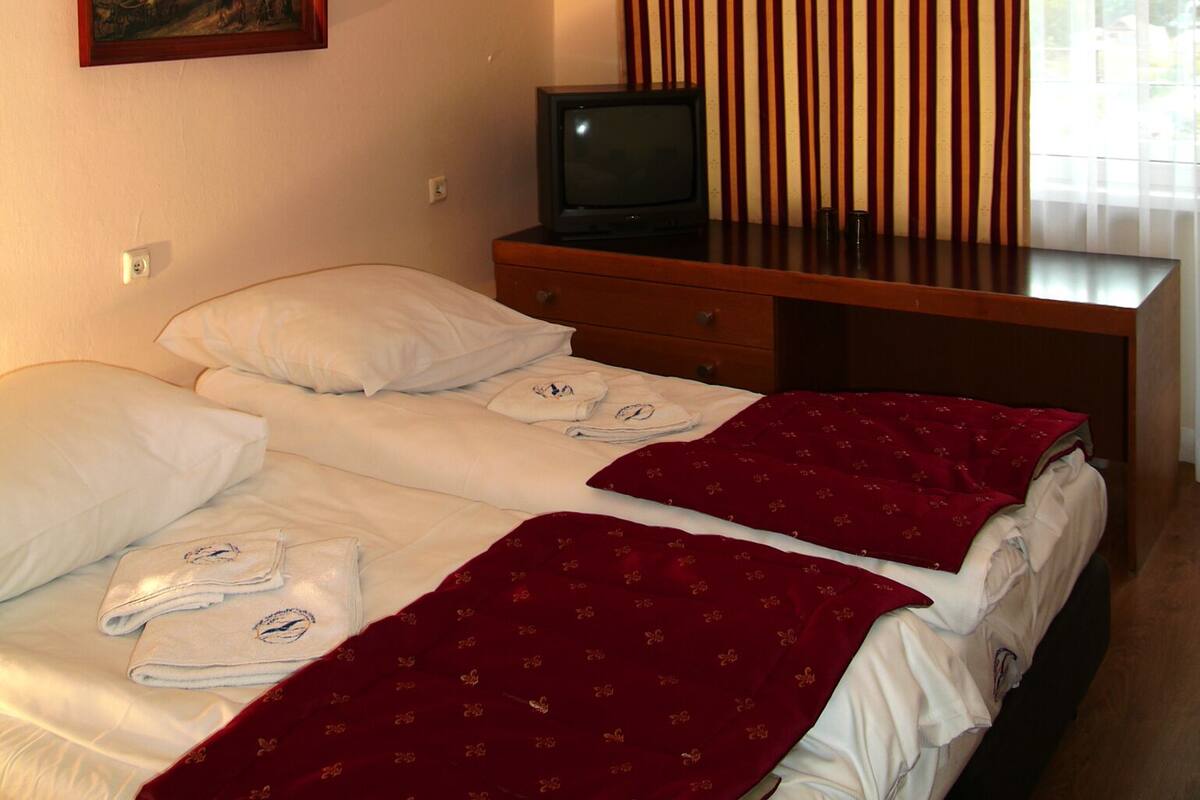 A hotel room with two beds, a television on a wooden dresser, a painting on the wall, and a window with vertical blinds.