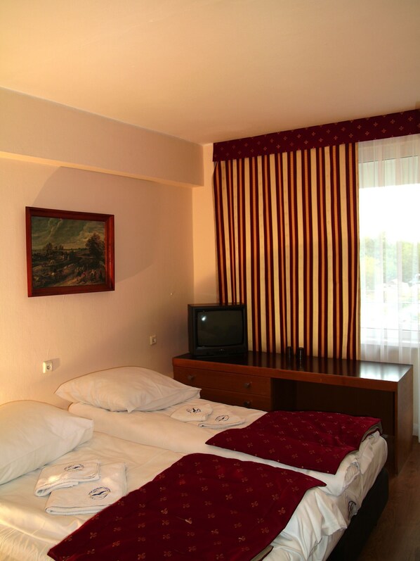 A hotel room with two beds, a television on a wooden dresser, a painting on the wall, and a window with vertical blinds.