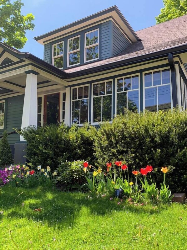 Une maison à deux étages dotée d’un porche, de grandes fenêtres et d’un jardin bien entretenu où poussent différentes sortes de fleurs.