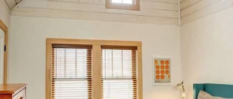 Un dormitorio con techo inclinado, vigas de madera, una cama con colchón azul, una mesita de noche de madera, una pequeña mesa con lámpara y una ventana con persianas.