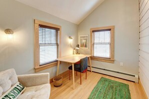 A small, cozy room with a wooden desk, two windows with blinds, a single chair, a couch, and a green rug.