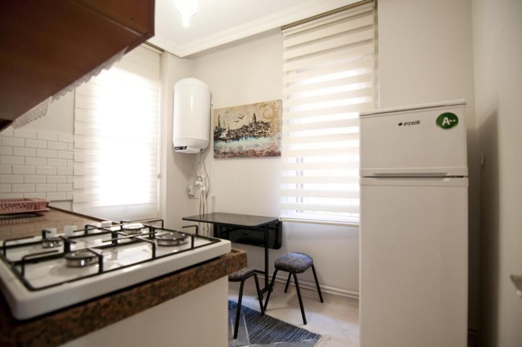 A kitchen with a gas stove, a small table, a stool, a refrigerator, and a painting on the wall.