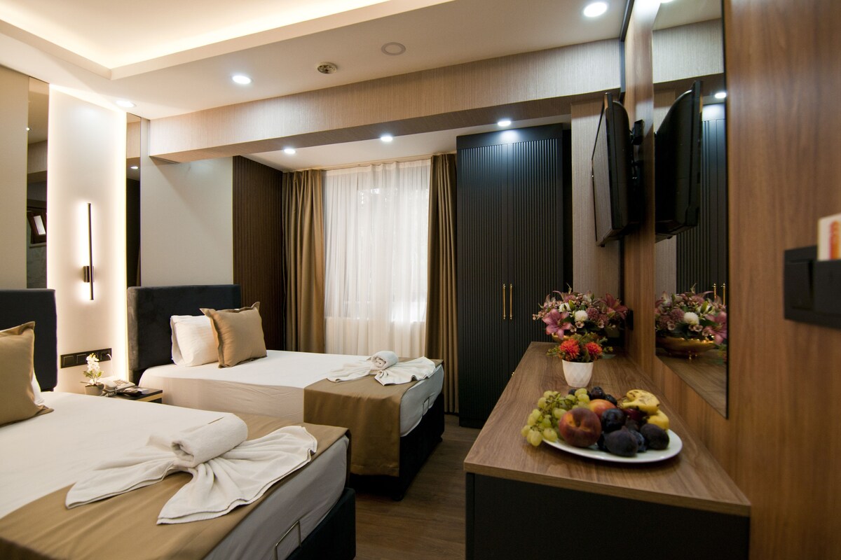 A hotel room with two beds, a wooden desk with a fruit plate, a mirror, and a TV on the wall.