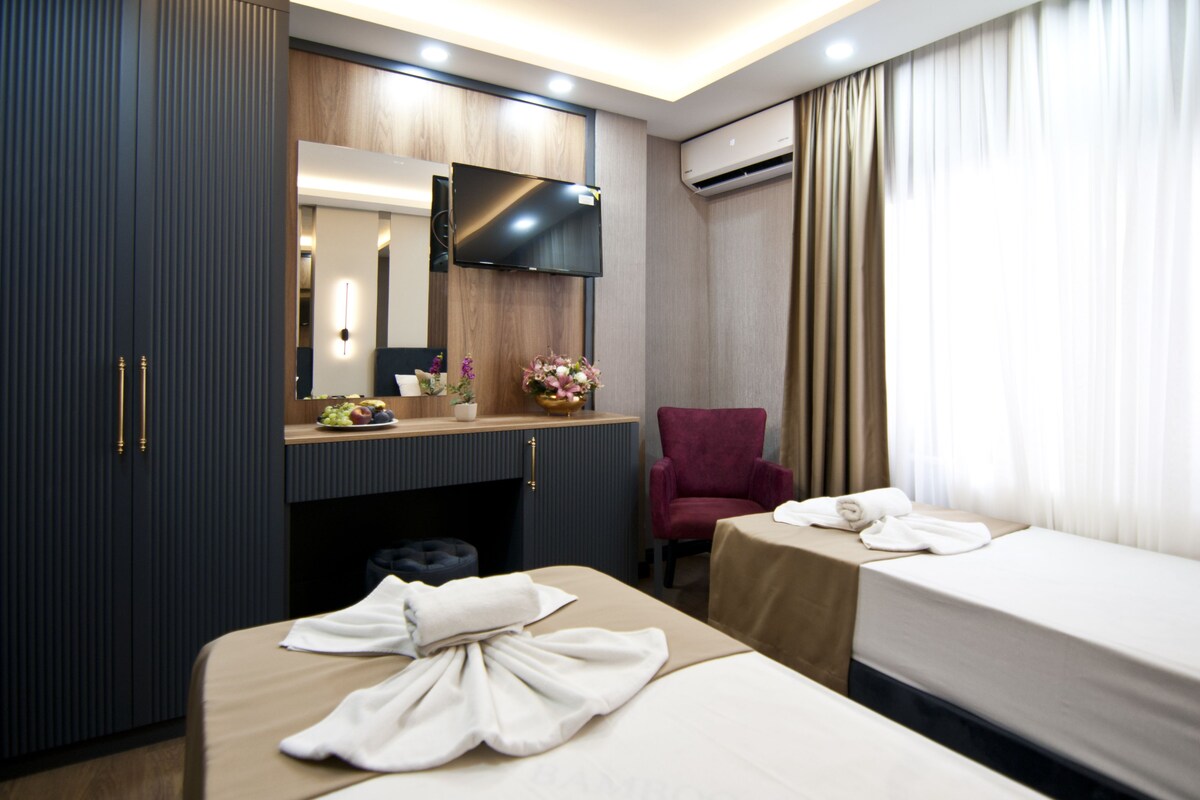 A hotel room with a bed, a desk with a flat-screen TV, a chair, and a small table with towels.