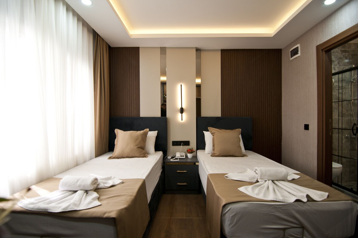 A hotel room with two beds, a nightstand, a wall-mounted light, and a window with curtains.