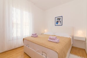 Un lit bien fait, recouvert d’une couverture beige, avec deux oreillers et deux serviettes pliées. Un tableau encadré est accroché au mur, au-dessus du lit.