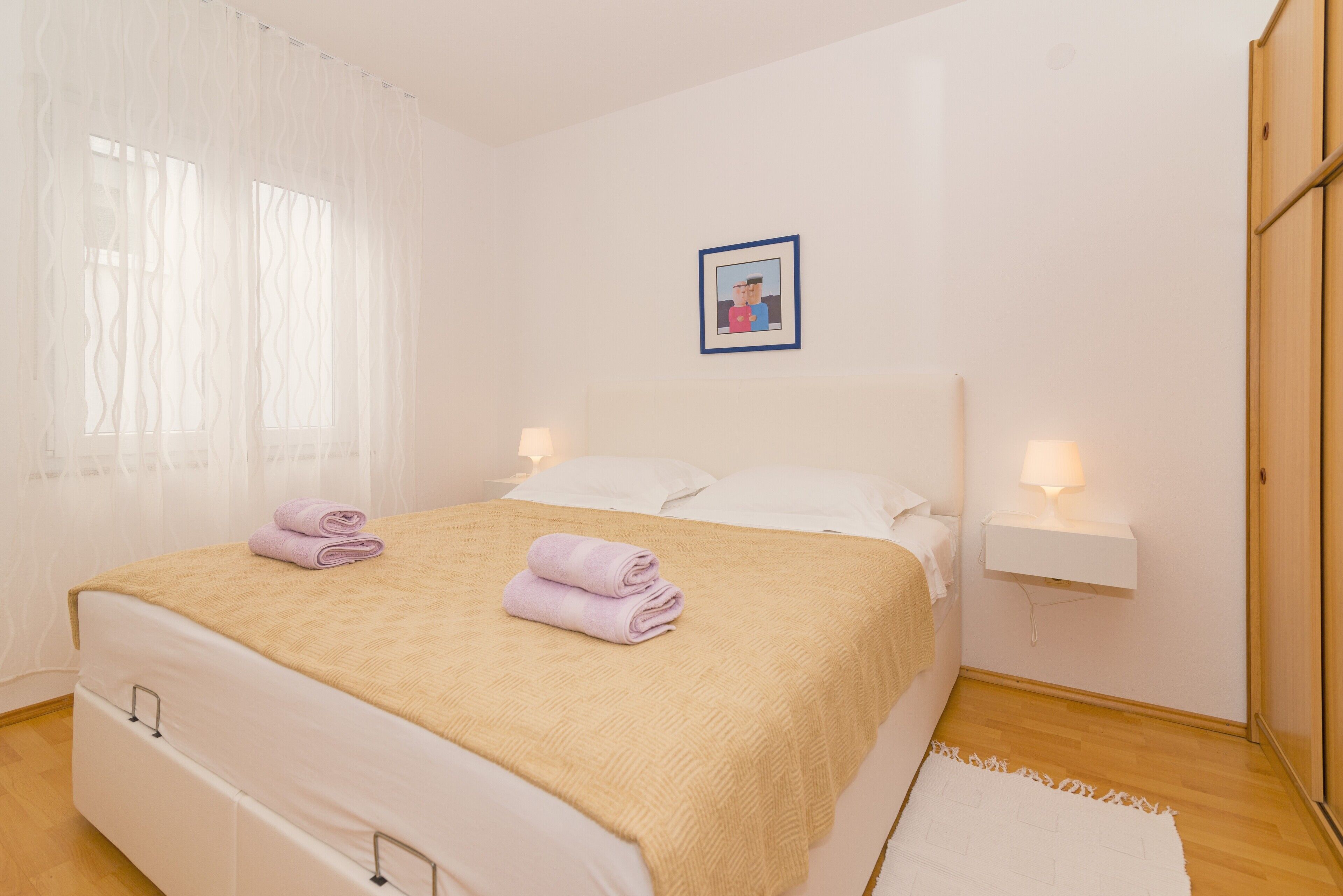 A neatly made bed with a beige blanket and two folded towels, a bedside table with a lamp, and a framed picture on the wall.