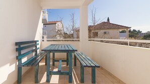 Ein Balkon mit einem Holztisch, zwei Bänken, einem Fliesenboden und Blick auf Gebäude und Bäume.