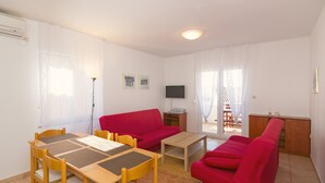 Ett vardagsrum med en röd soffa, ett träbord och ett matbord med stolar. Det finns en tv på väggen och ett fönster med gardiner.