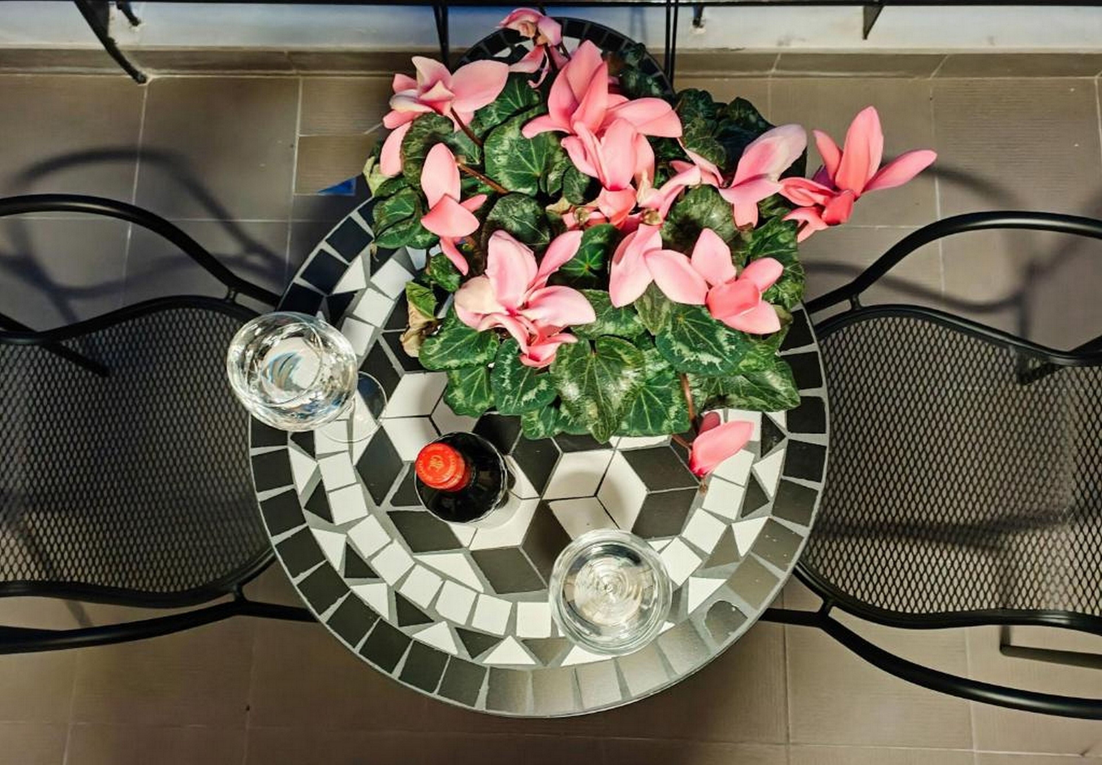 A round table with a mosaic tile design, two chairs, a potted plant with pink flowers, and two glasses.