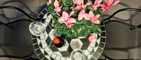A round table with a mosaic tile design, two chairs, a potted plant with pink flowers, and two glasses.