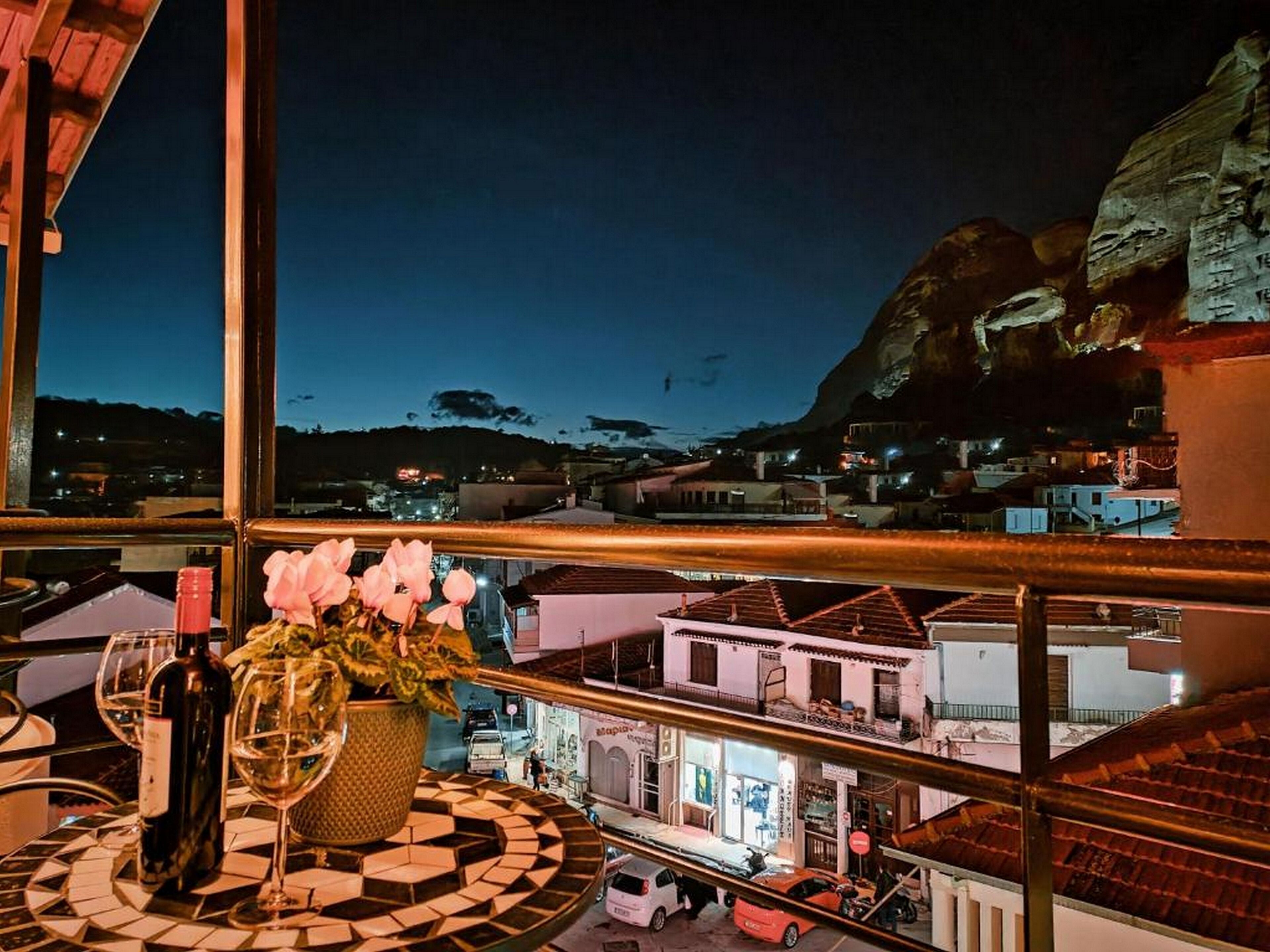 A balcony with a view of a town at dusk, a table with a bottle and glasses, and a vase with flowers.
