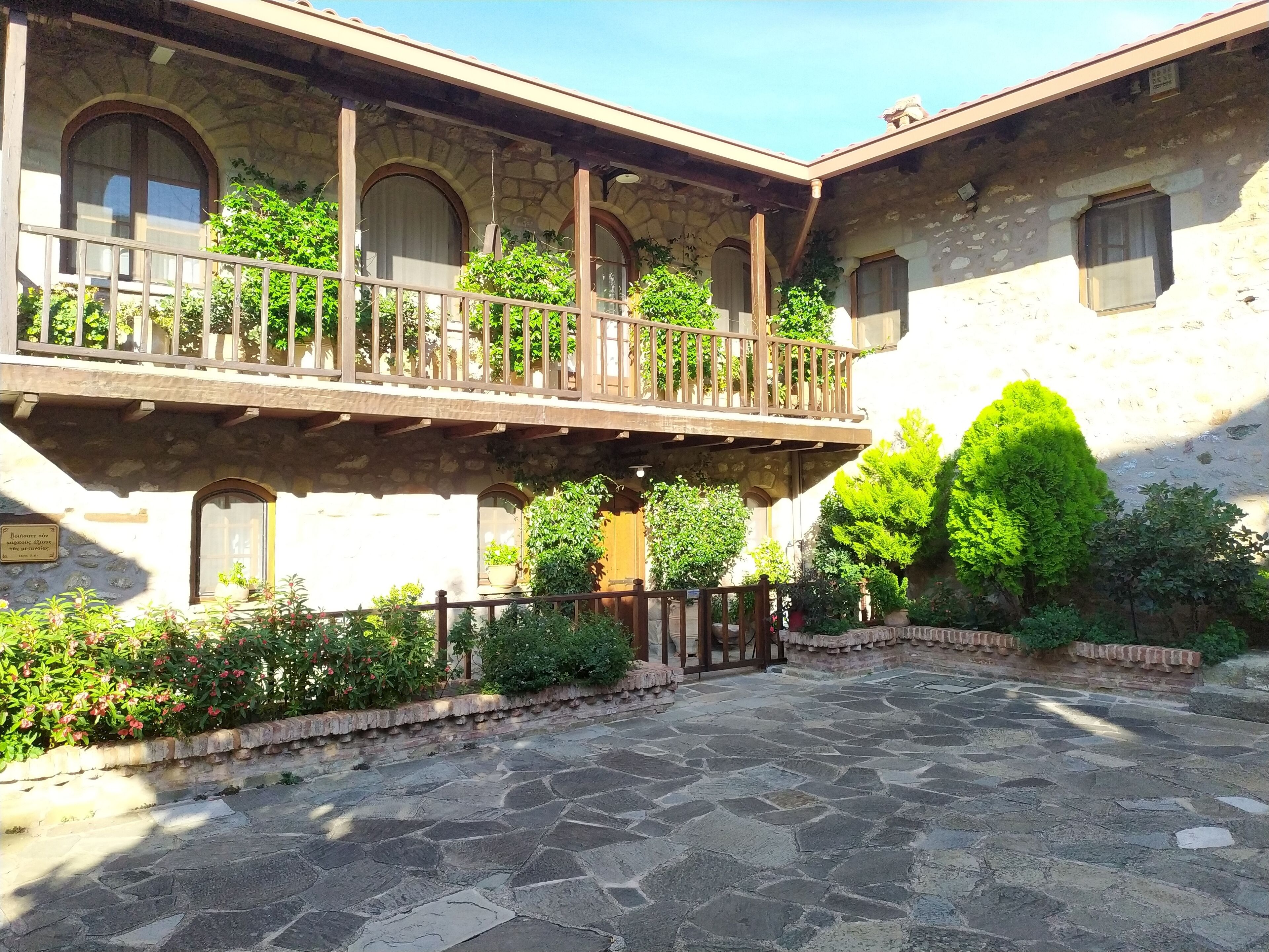 Une cour dallée de pierres, un balcon avec des balustrades en bois et une végétation luxuriante.