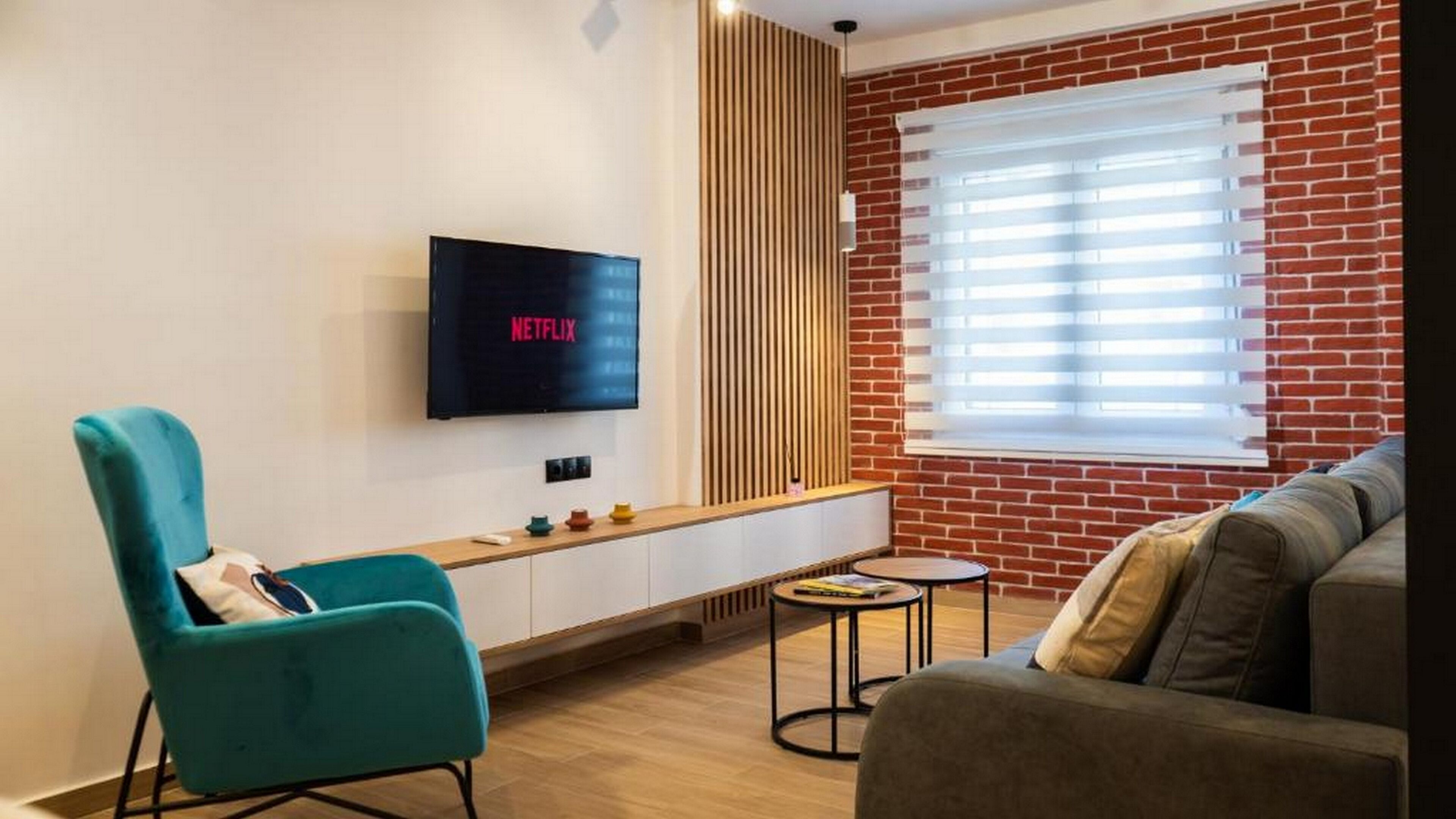 A modern living room with a flat-screen TV displaying 'NETFLIX', a teal armchair, a grey sofa, a small round coffee table, and a brick wall with a window with blinds.