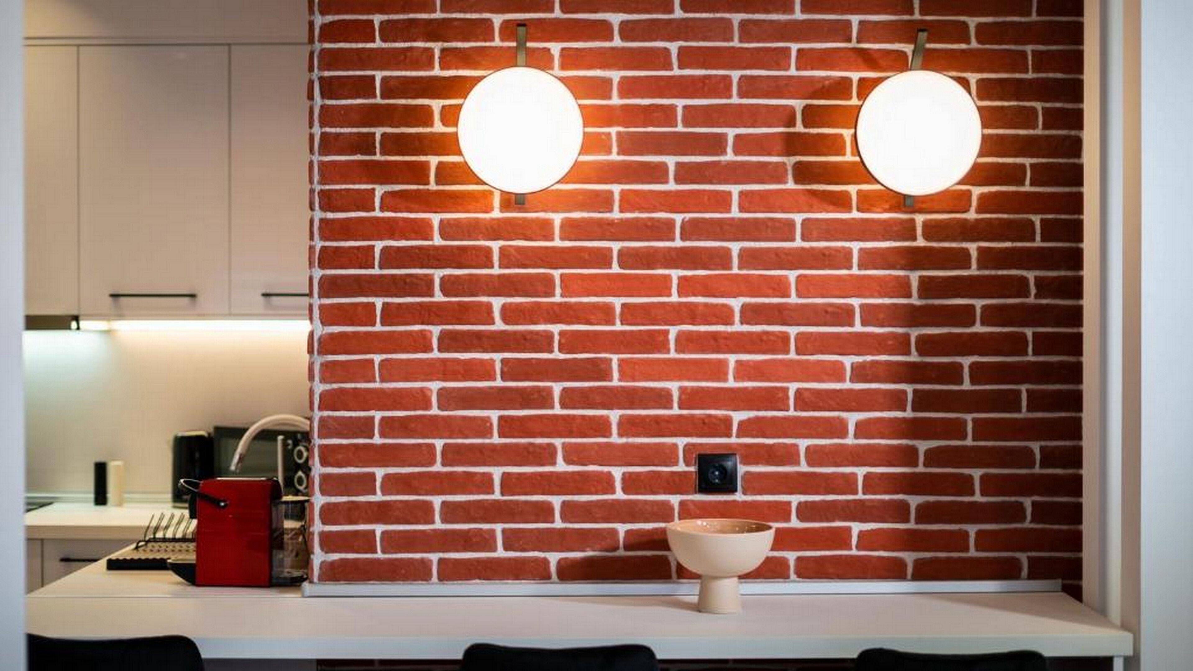 A kitchen with a brick wall, two round wall-mounted lights, a white countertop, and a small beige bowl.