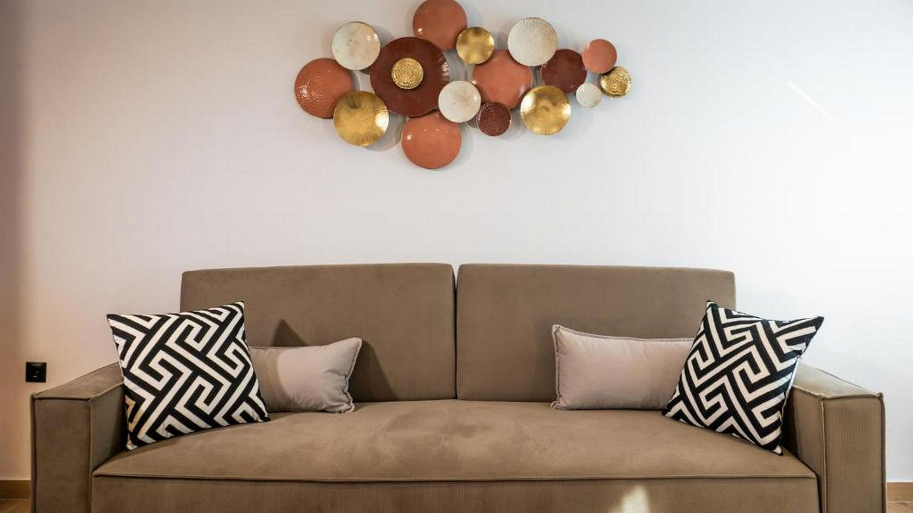 A beige sofa with two patterned pillows, a wall-mounted abstract art piece, and a plain wall.