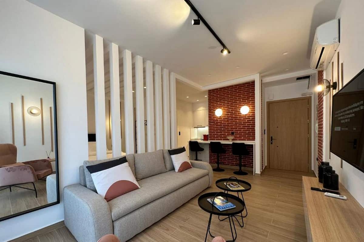 A modern living room with a sofa, coffee table, and chairs. There is a kitchen area with a brick wall and bar stools.