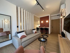 Uma sala de estar moderna com sofá, mesa de centro e cadeiras. Há uma área de cozinha com parede de tijolos e banquetas.