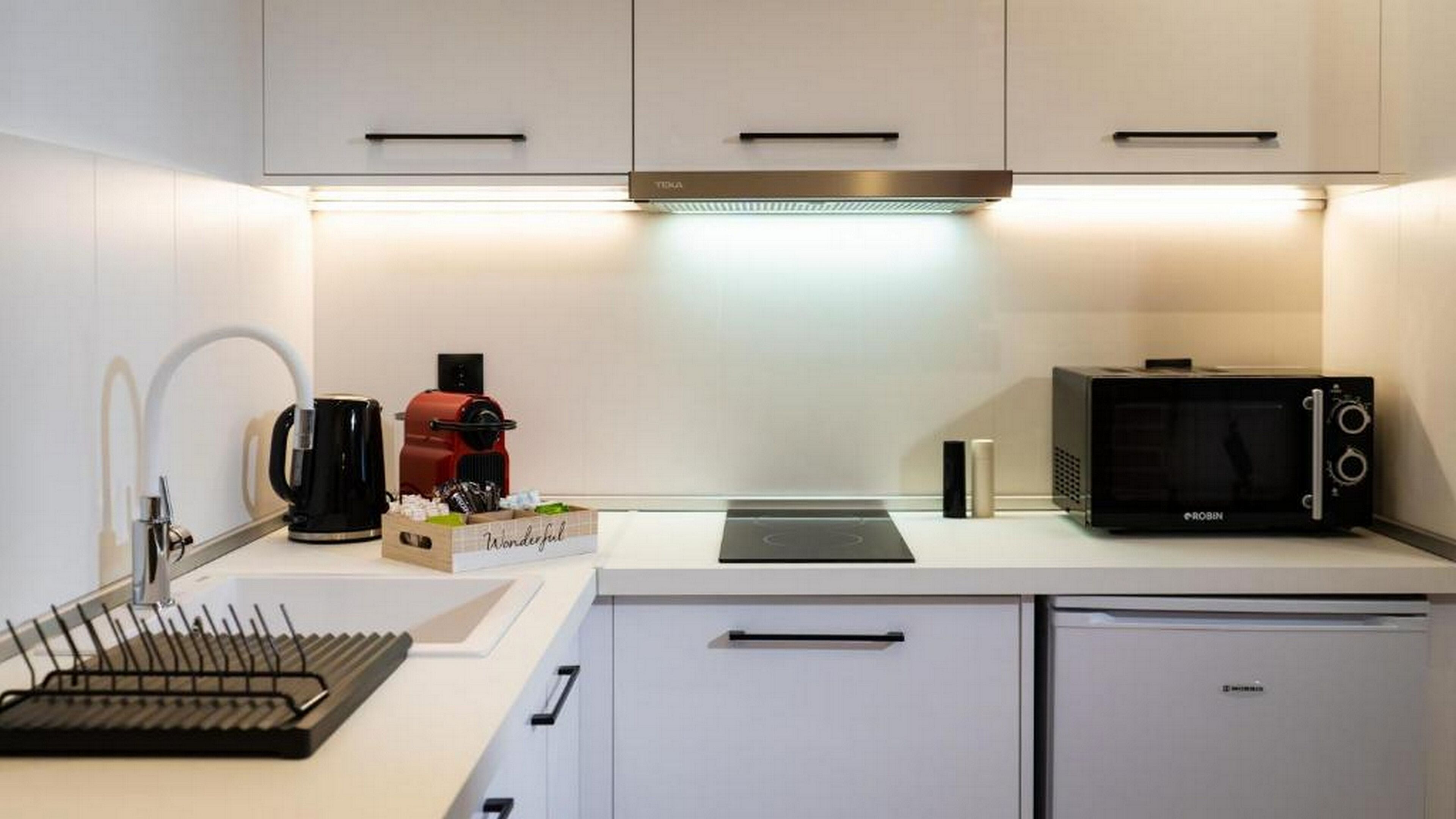 A modern kitchen with white cabinetry, a black coffee machine, a red coffee grinder, a black microwave, and a small refrigerator.