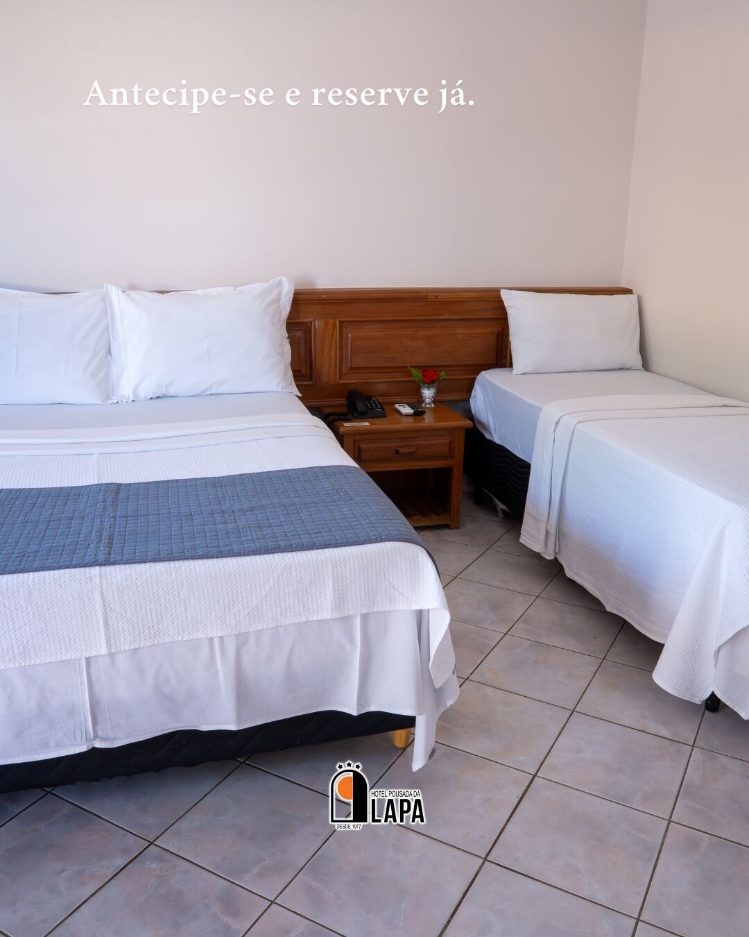 A hotel room with two beds, a wooden headboard, a nightstand with a phone and remote, and a tiled floor.
