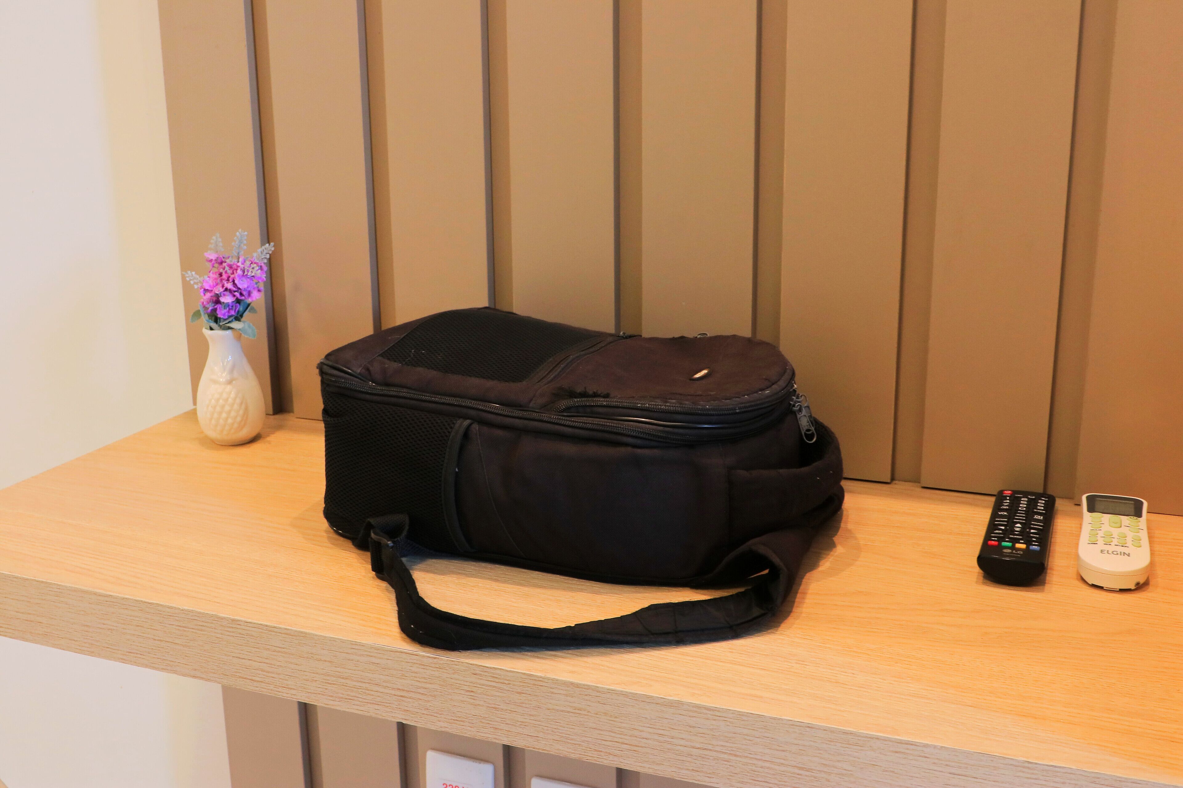 A black bag on a wooden surface with a vase of flowers and two remote controls.