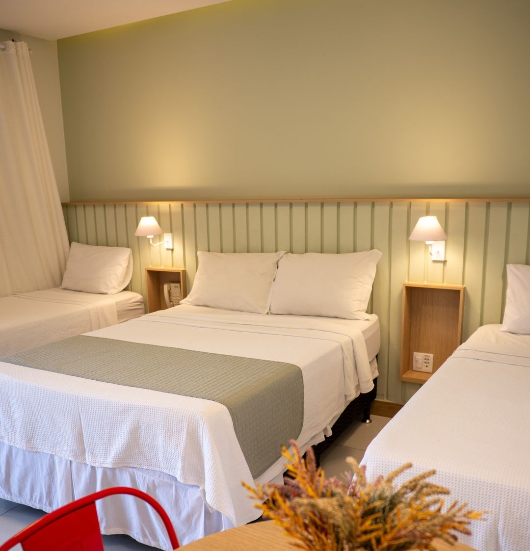 Habitación de hotel con dos camas, una mesita de noche de madera, una silla roja y un jarrón con flores secas.