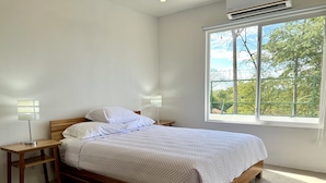 Un letto rifatto con cura, con un telaio in legno e biancheria bianca, due comodini con lampade e un'ampia finestra vista alberi e recinzione.
