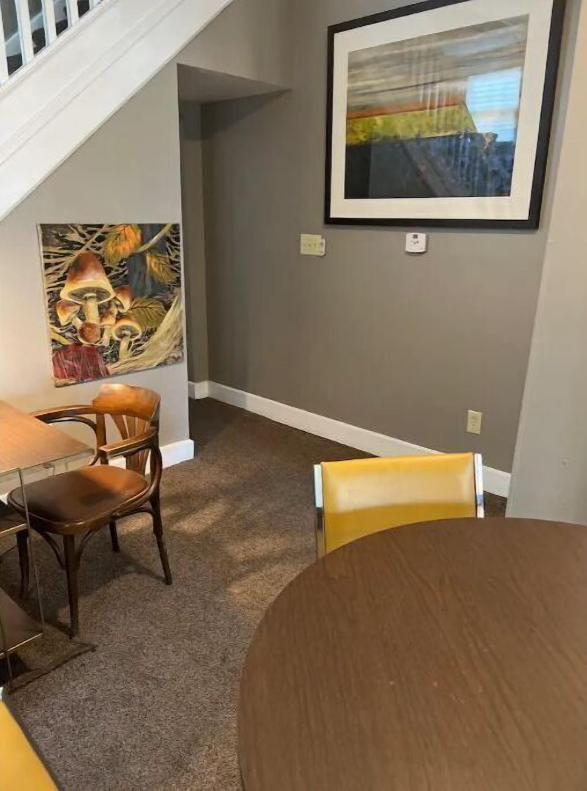 A dining area with a round table and two chairs, a painting on the wall, and a staircase in the background.