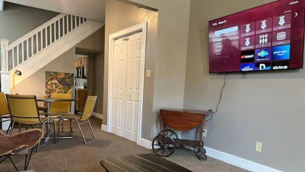 A living room with a TV mounted on the wall, a dining area with yellow chairs, and a staircase in the background. - IU Medical Campus Convenience (Indianapolis)