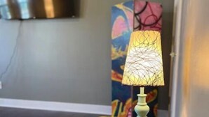 A yellow armchair and a table lamp in a room with a flat-screen TV mounted on the wall and a colorful abstract painting in the background. - IU Medical Cozy Campus Castle (Indianapolis)