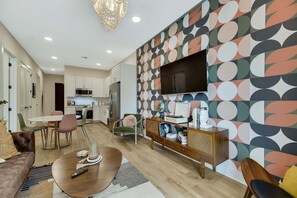A modern living room with a dining area, a kitchen in the background, and a TV mounted on a colorful wall.