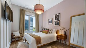 A modern bedroom with a large bed, a wooden nightstand, a mirror, and a view of the city through large windows.