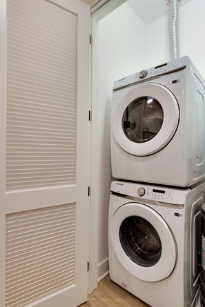 A laundry room with two stacked washing machines and a closed door with horizontal blinds.