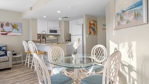 A modern kitchen with white cabinets, a microwave, and a refrigerator. There is a dining area with a glass table and white chairs. - Gulf Shores Plantation-West 1240 (Gulf Shores)