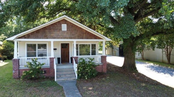 A single-story house with a front porch, brick foundation, and a large tree in the yard.