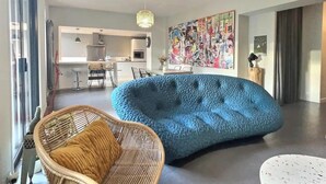 Un soggiorno moderno con un divano blu, poltrone in vimini e una cucina sullo sfondo.