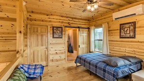 Quarto de cabana de madeira com cama, um banco, ventilador de teto e porta que dá para outro cômodo.