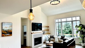 A spacious living room with a fireplace, a large window, and a modern chandelier. - New Luxury House, walk to Asheville’s best restaurants, pubs breweries and shops (Asheville)