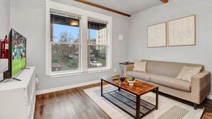 A living room with a beige sofa, a coffee table, and a TV on a stand. There are two windows with a view of the street and parked cars. - Walk to DT | Fast Wi-fi + Long stay equipped! (Tacoma)
