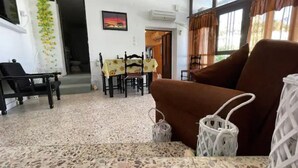A living room with a brown sofa, a dining table with chairs, a small table with a white basket, and a painting on the wall. - Nionas Cheerful House (Piso Meria)