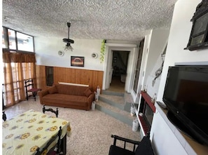 Un salon avec un canapé marron, une table à manger recouverte d’une nappe à motifs floraux, un ventilateur de plafond et un téléviseur.