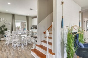 Un coin repas avec une table ronde et des chaises blanches, un escalier avec des marches en bois et une rampe blanche, et une chaise bleue agrémentée de grandes plantes vertes.