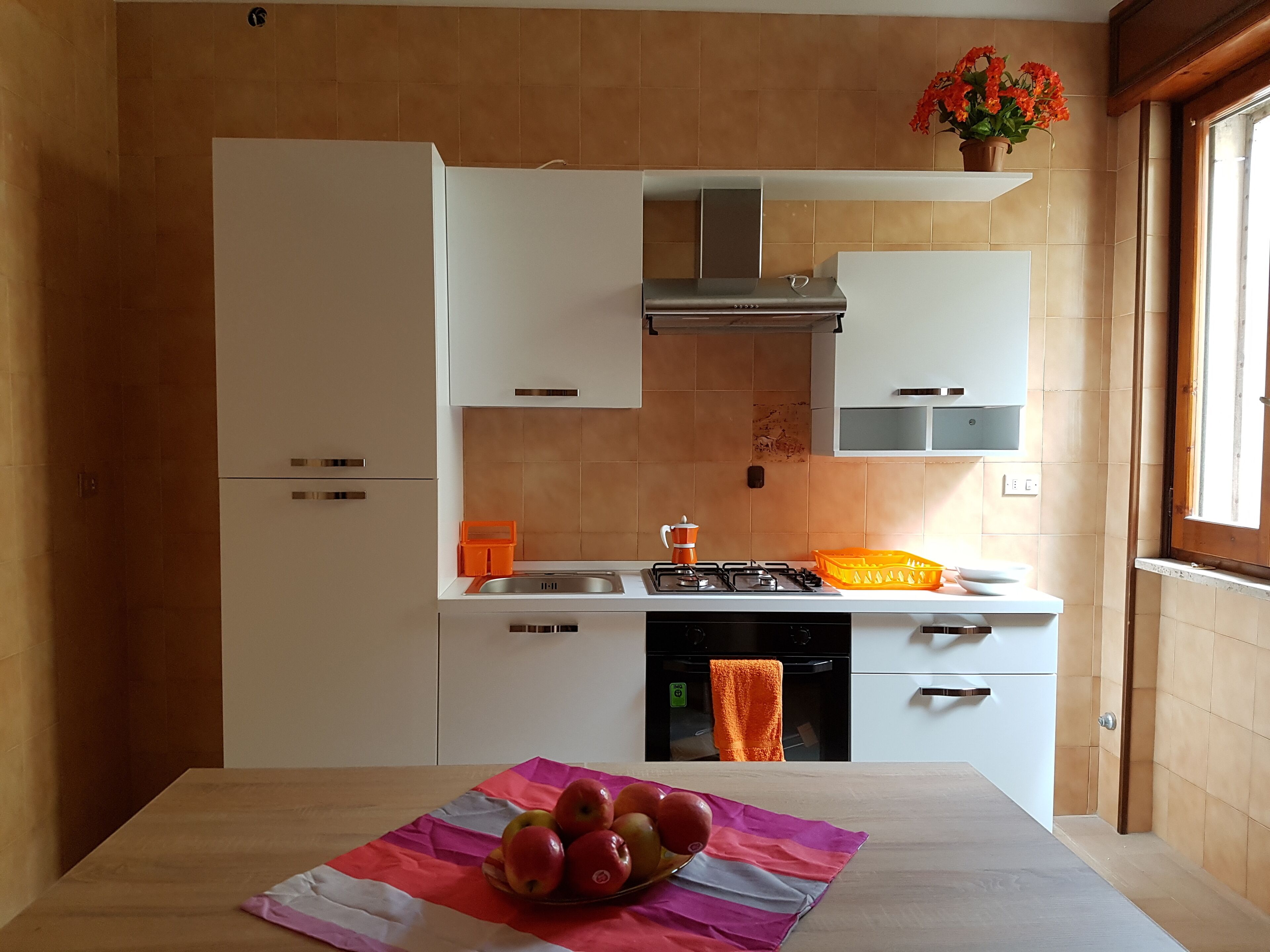 Una cucina moderna con mobili bianchi, un fornello in acciaio inossidabile e una finestra con fiori arancioni.