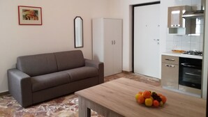 A modern living room with a grey sofa, a wooden coffee table with fruit, a white wardrobe, and a kitchen with a stove and oven. - Lido San Giovanni (Gallipoli)