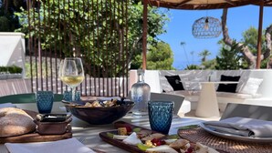Un espace de restauration en plein air avec une table dressée pour un repas, comprenant du pain, un bol d’olives et une bouteille de vin.