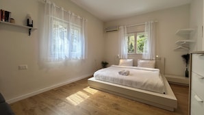 Un dormitorio con cama, dos ventanas con cortinas translúcidas, una mesita de noche con lámpara y una pequeña estantería con libros.