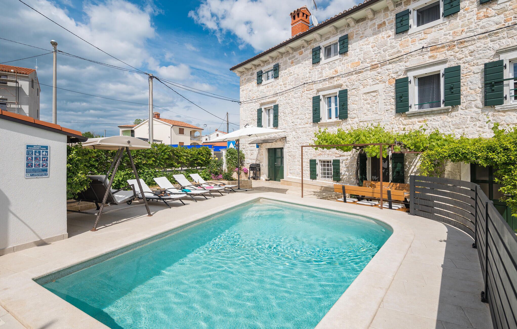A swimming pool area with lounge chairs, a stone building with green shutters, and a sign indicating a swimming pool.