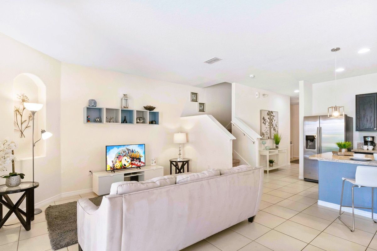 Una sala de estar moderna con sofá, televisor y objetos decorativos. Se ve una cocina con refrigerador y una isla en el fondo.